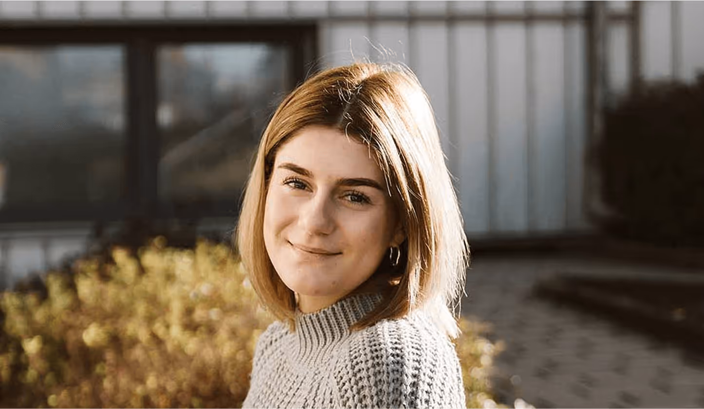 Frau mit kurzem, blondem Haar lächelt in die Kamera, trägt grauen Pullover, im Hintergrund unscharfe Pflanzen und Gebäude.