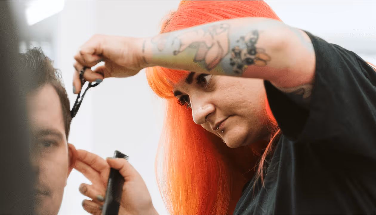 Friseurin mit rotem Haar und Tattoos schneidet konzentriert die Haare eines Kunden in einem modernen Salon.
