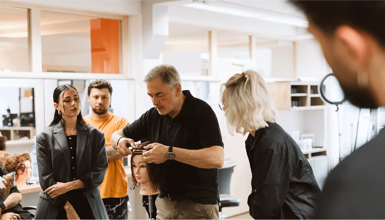 Frisörkurs: Mann schneidet Haarpuppe, andere Teilnehmer beobachten konzentriert im Salon.