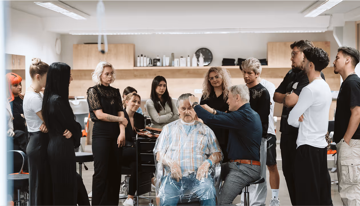 Friseur lehrt Haarschneidetechniken umringt von Lehrlingen, während älterer Mann auf Stuhl sitzt. Training in Friseursalon.