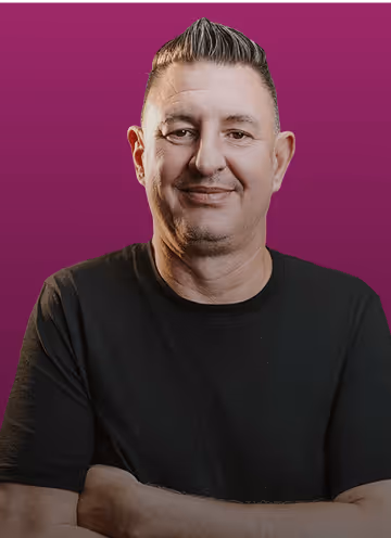 Mann mit dunklem T-Shirt und kurzen Haaren vor lila Hintergrund, lächelnd mit verschränkten Armen.