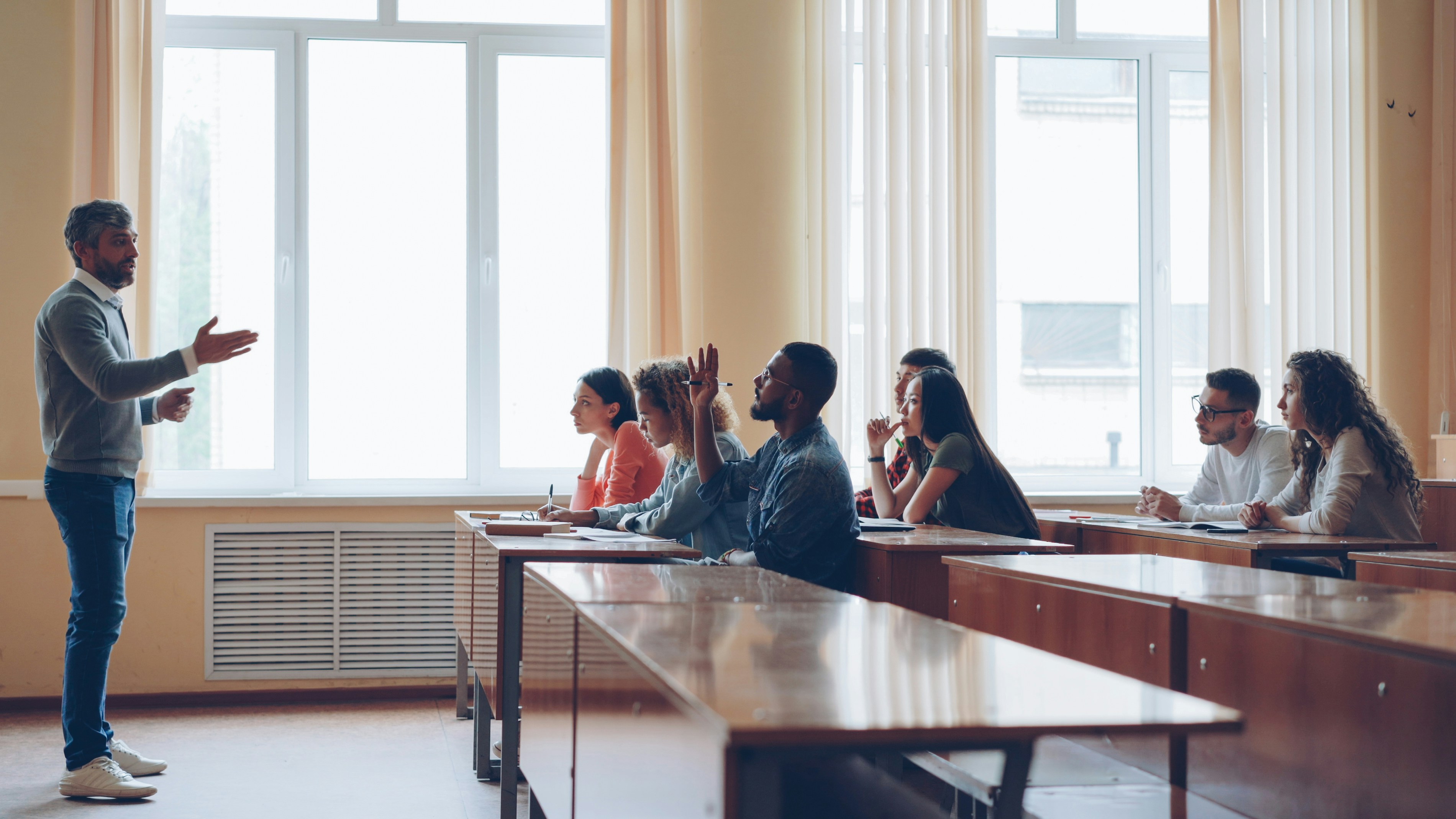 Teacher speaking to attentive students seated in a classroom with large windows behind them.