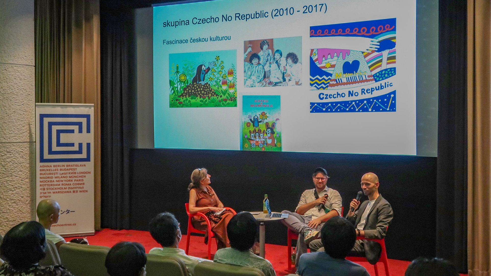 Three speakers seated on stage with a projected slide about Czecho No Republic and Czech culture images.