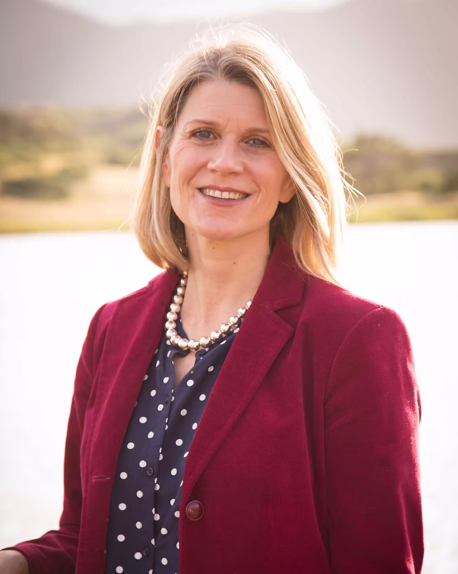 Smiling woman with blonde hair wearing a burgundy blazer, navy polka dot blouse, and pearl necklace, standing outdoors by water.