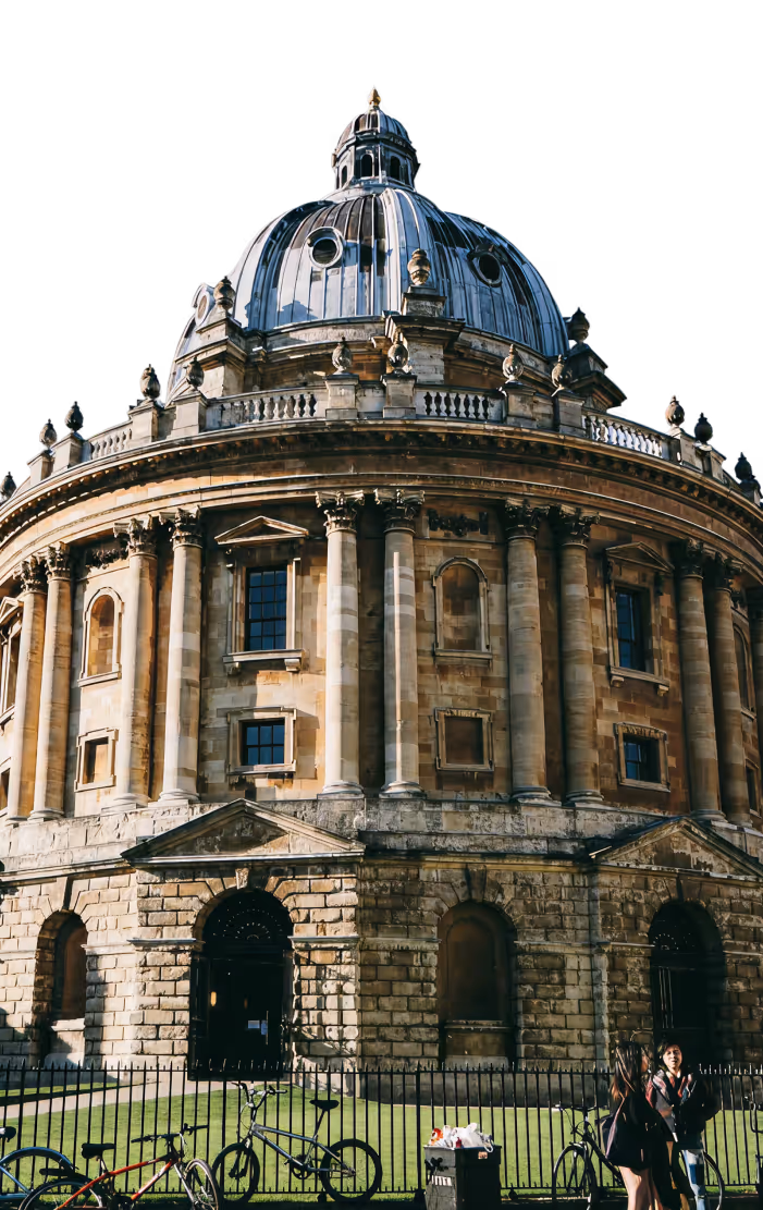A prominent, eye-level view of the Radcliffe Camera in Oxford, a large circular building in the English Baroque style, featuring a high, dark lead-covered dome. The building's stone exterior is richly detailed with columns and windows. To the left, a portion of the older, darker, Gothic-style architecture of another university building is visible. The sky is bright, almost white, highlighting the building's texture.