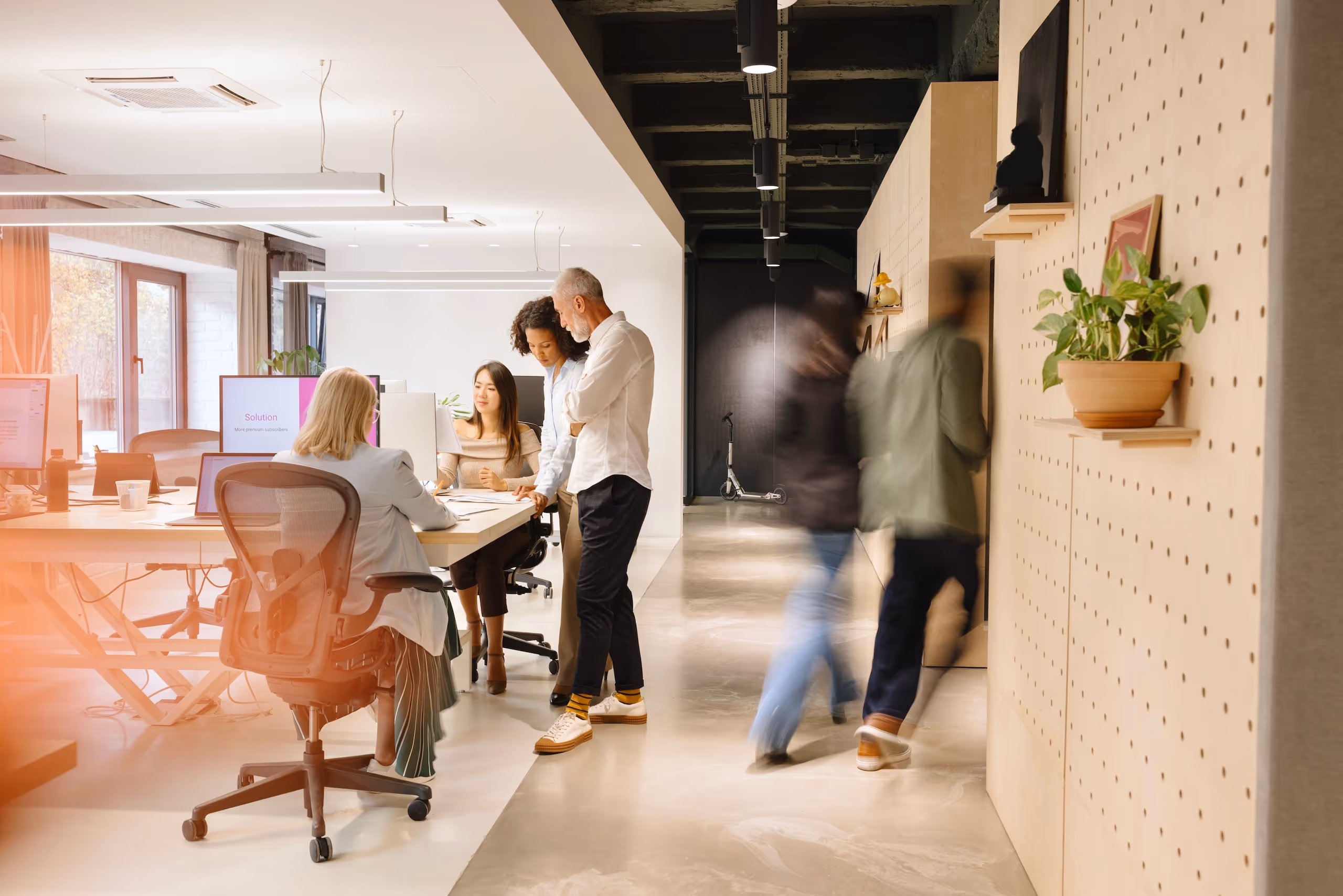 Group of diverse coworkers collaborating at a modern white office desk with computers while two blurred figures walk in the background.