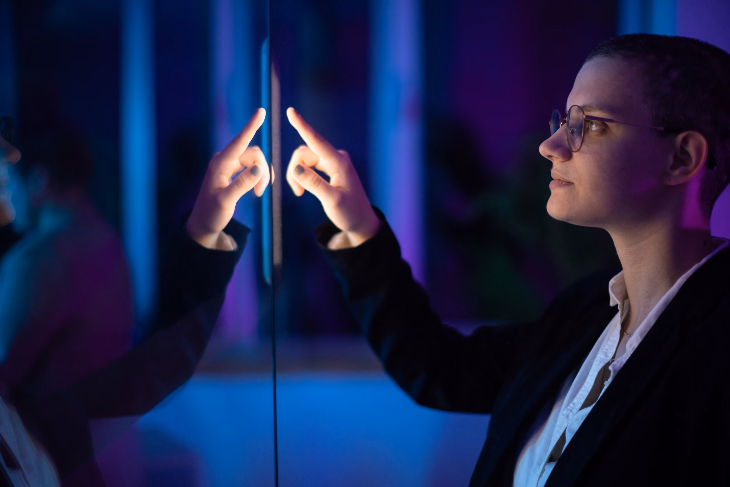 Person with short hair and glasses touching a transparent screen illuminated with blue and purple light.