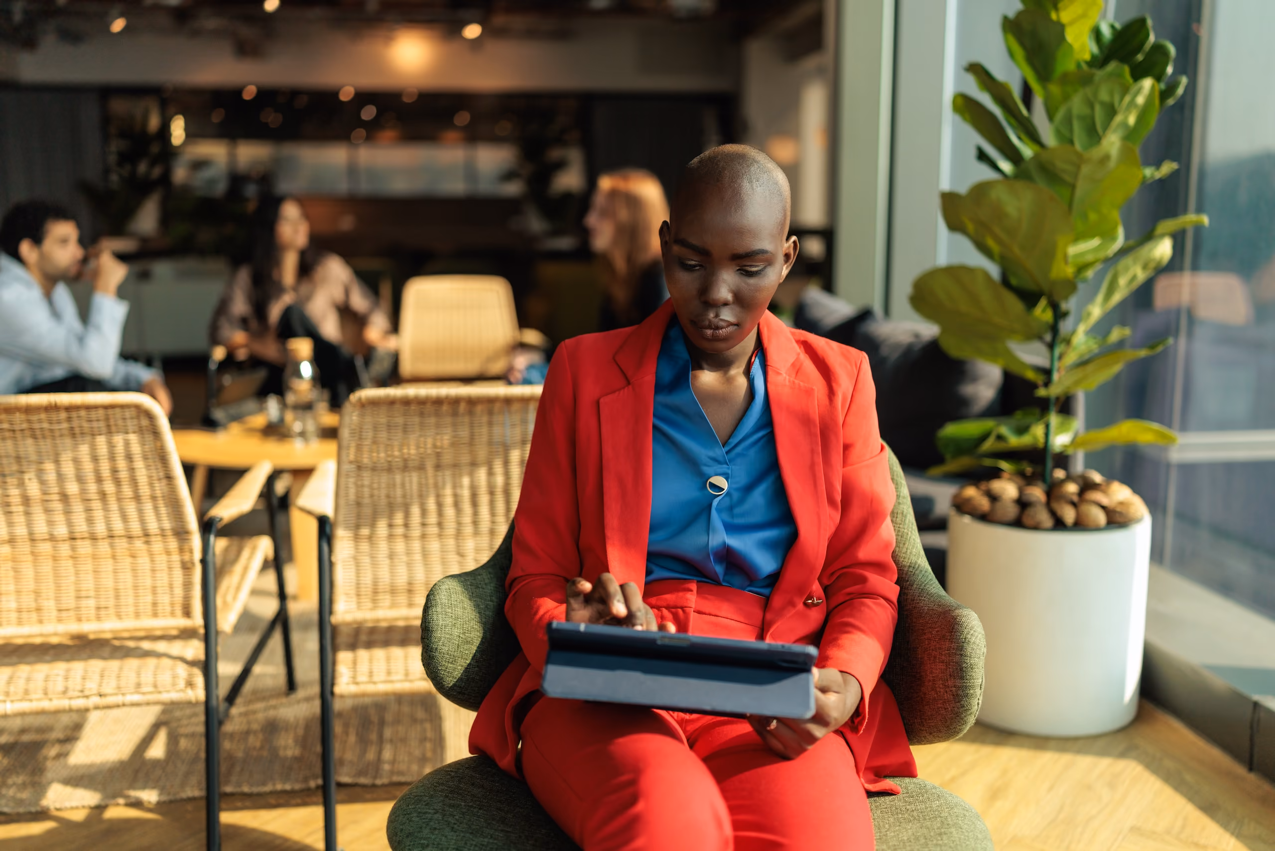 A woman in a red suit and blue blouse sits in a green chair using a tablet in a modern office with three blurred people and plants in the background.