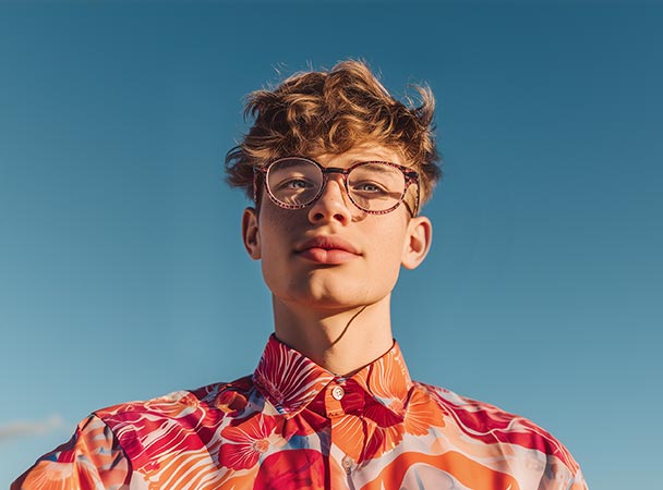 Young person wearing round glasses and a bright floral shirt, looking confidently at the camera against a clear blue sky