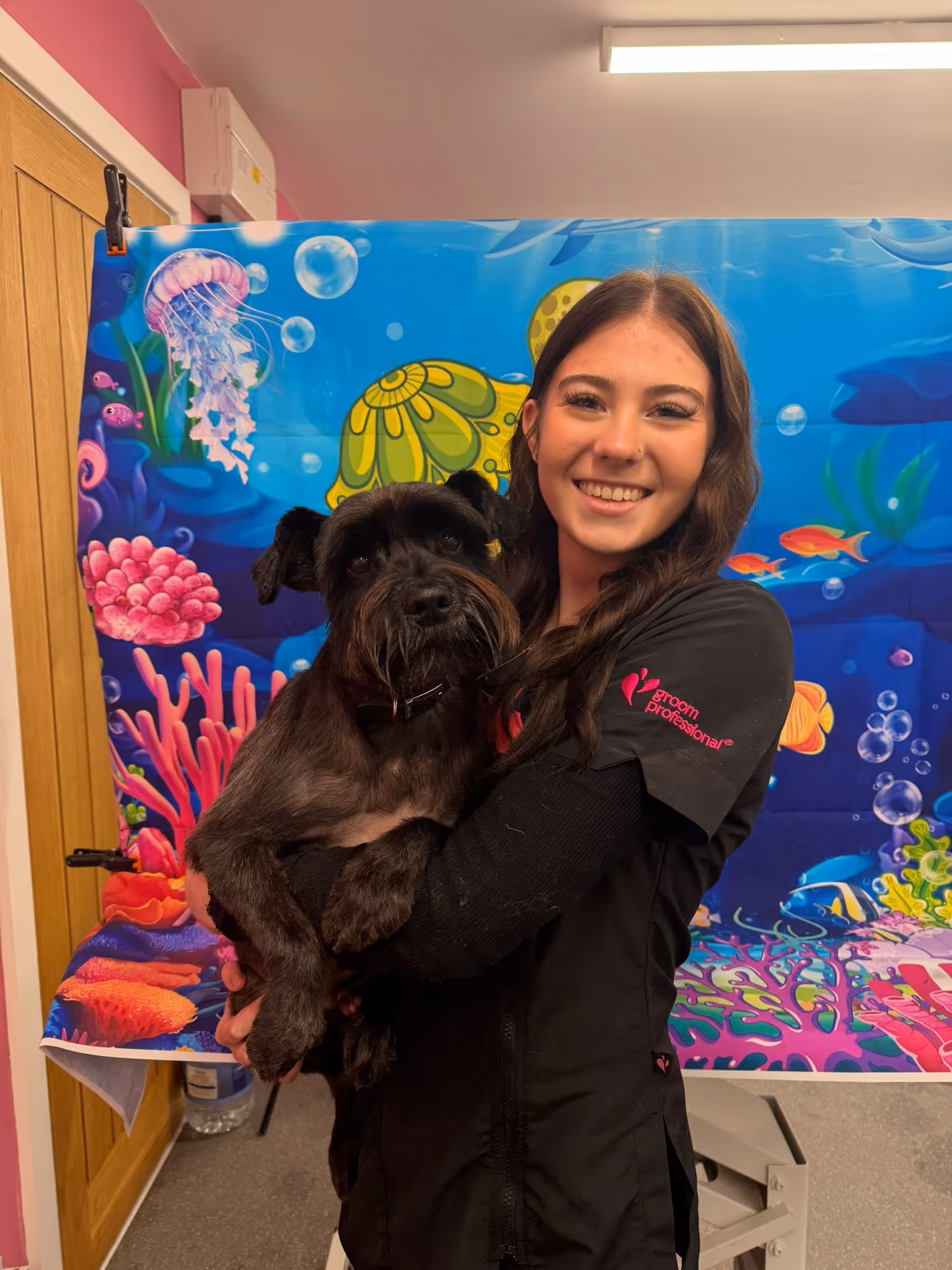 Smiling woman in black grooming uniform holding a freshly groomed black dog in front of a colorful underwater-themed backdrop.