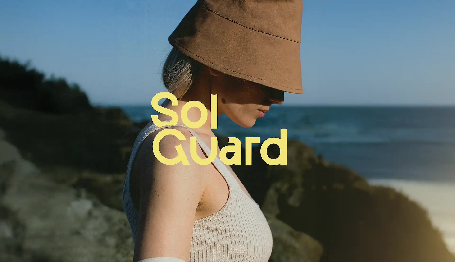 Woman wearing a beige bucket hat and sleeveless top standing by rocky shore with ocean in the background.