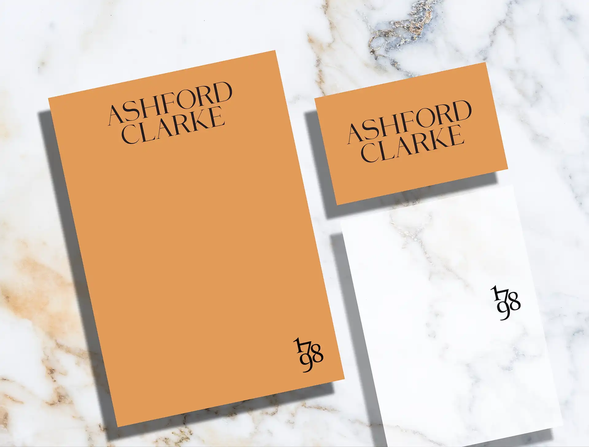 Stationery set on marble surface featuring orange card and folder with 'Ashford Clarke' in black serif font and black '1798' emblem on the folder and white envelope.