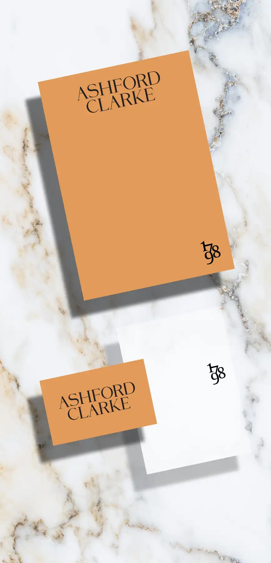 Two Ashford Clarke branded cards, one orange with black text and a date ‘1798’, and one white with the same date, placed on marble surface.