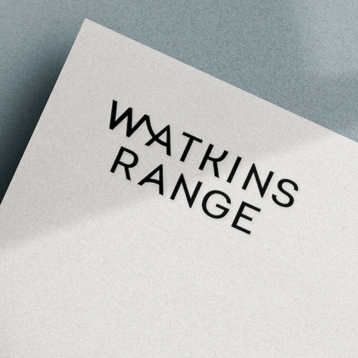 Close-up of a white card with the black text 'WATKINS RANGE' on a textured blue-grey surface.