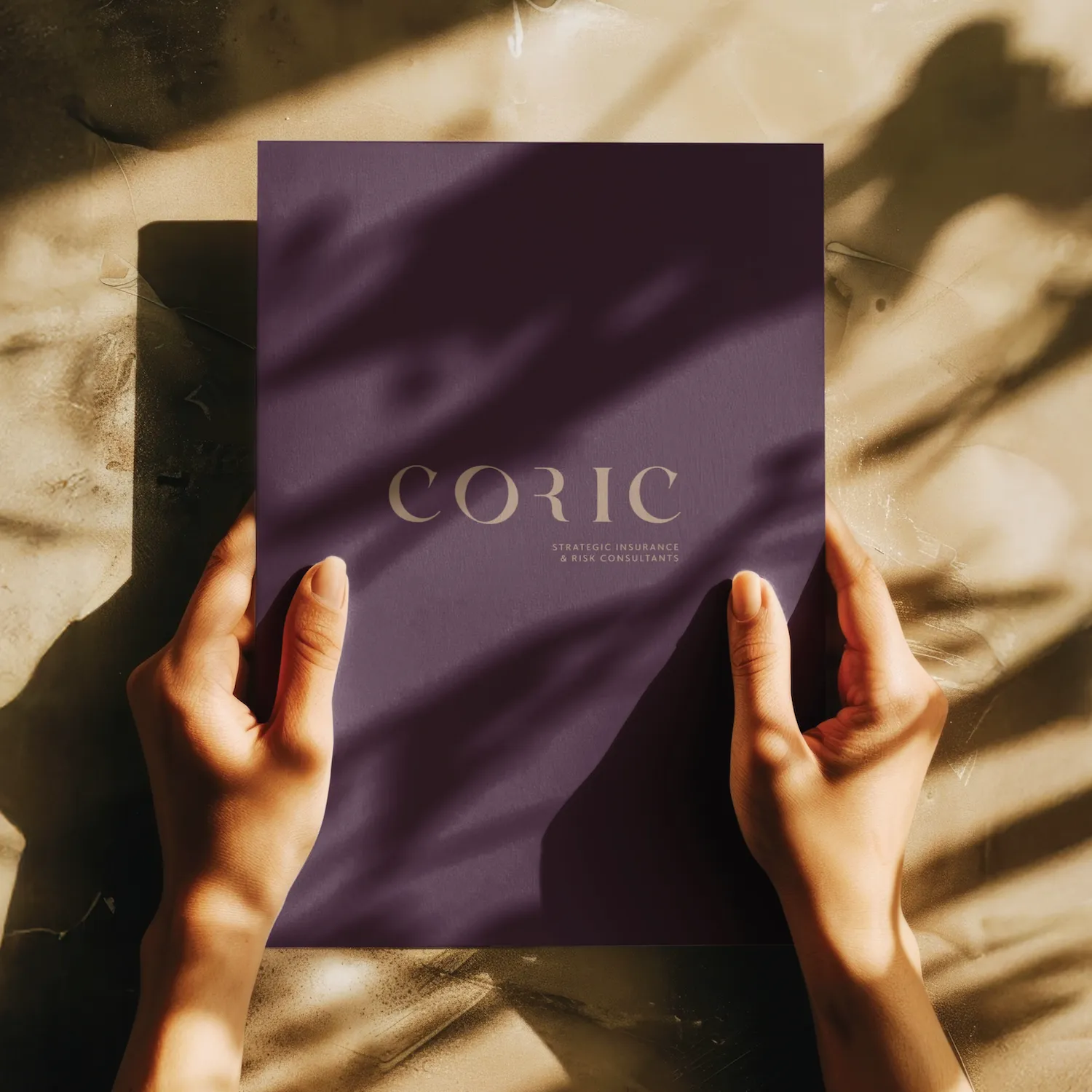 Two hands holding a dark purple folder with the text 'CORIC Strategic Insurance & Risk Consultants' on a textured beige surface with shadows.