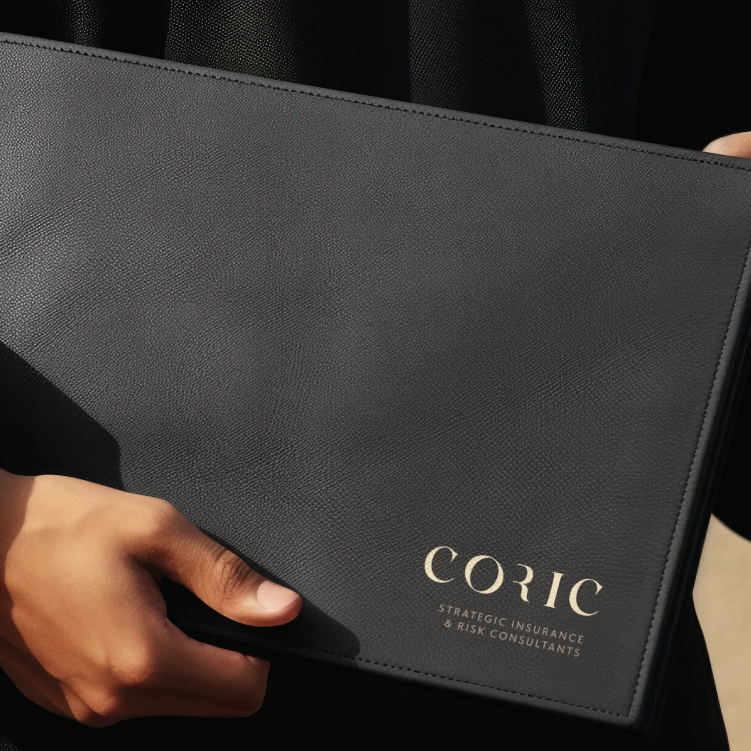 Close-up of a hand holding a black leather folder embossed with the text 'CORIC Strategic Insurance & Risk Consultants’.