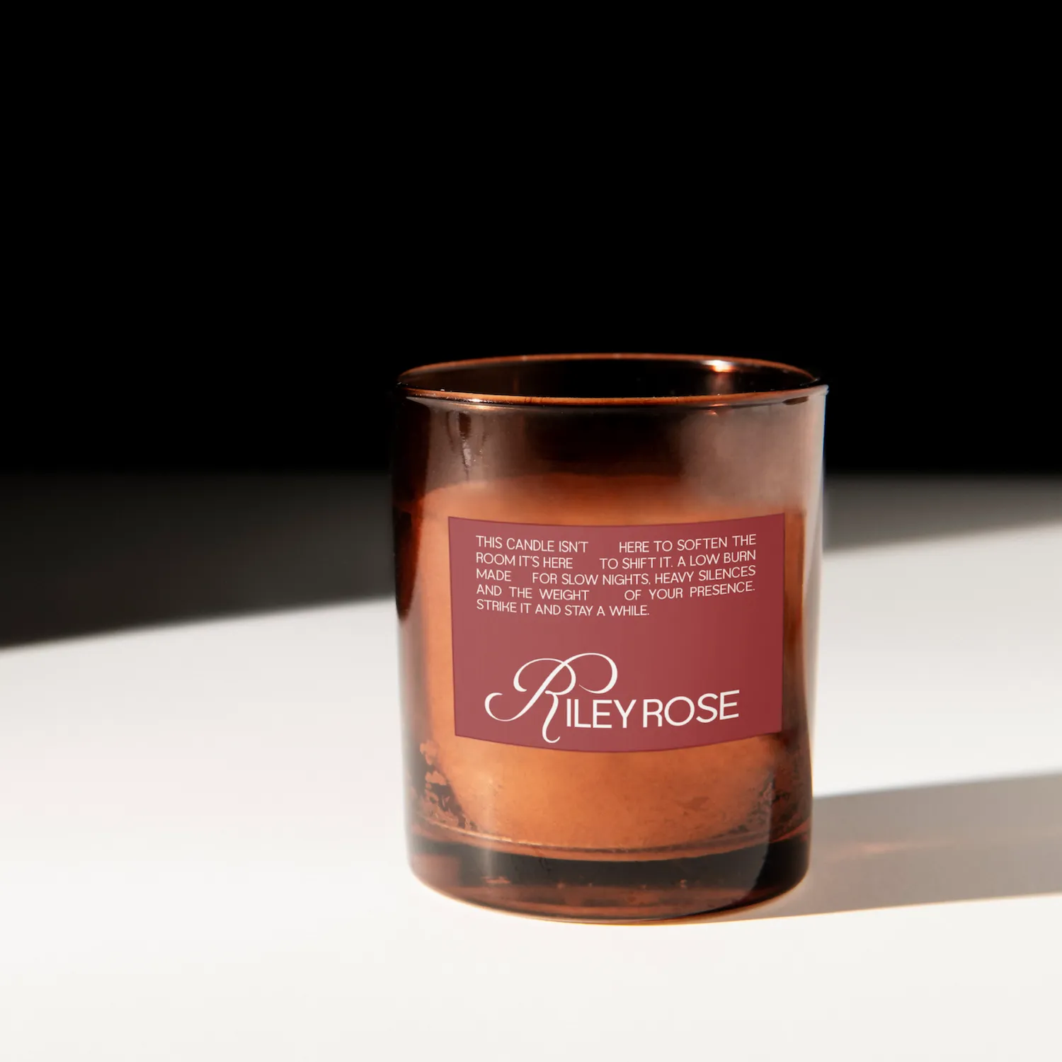 Brown glass candle holder with a maroon label reading 'Riley Rose' and a poetic description about the candle.