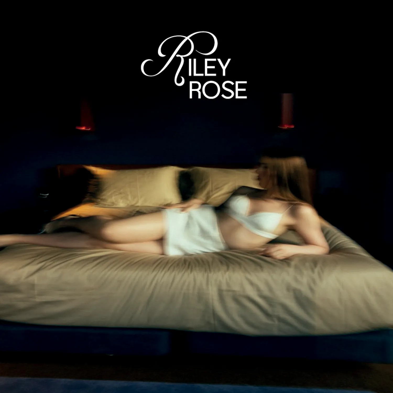 Woman in white lingerie reclining on beige bed with dark background and Riley Rose logo above.