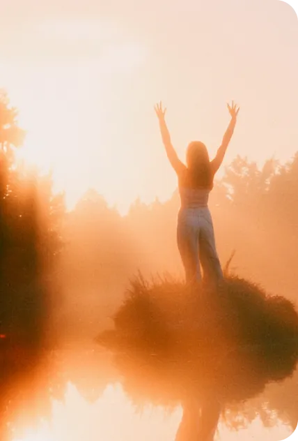 Woman raising arms in golden sunrise mist.