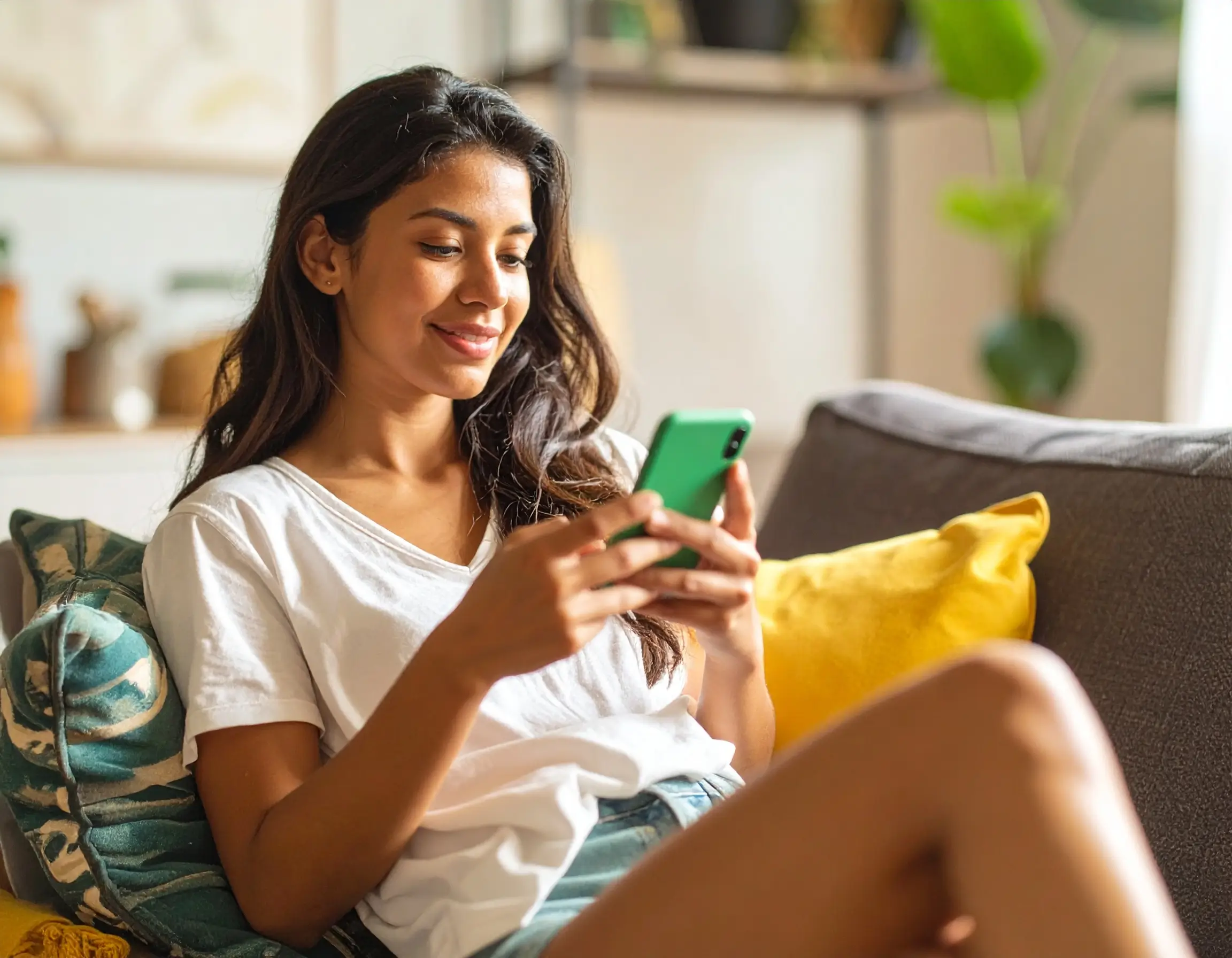 Foto de uma mulher jovem sentada em um sofá com almofadas coloridas sorrindo para seu celular