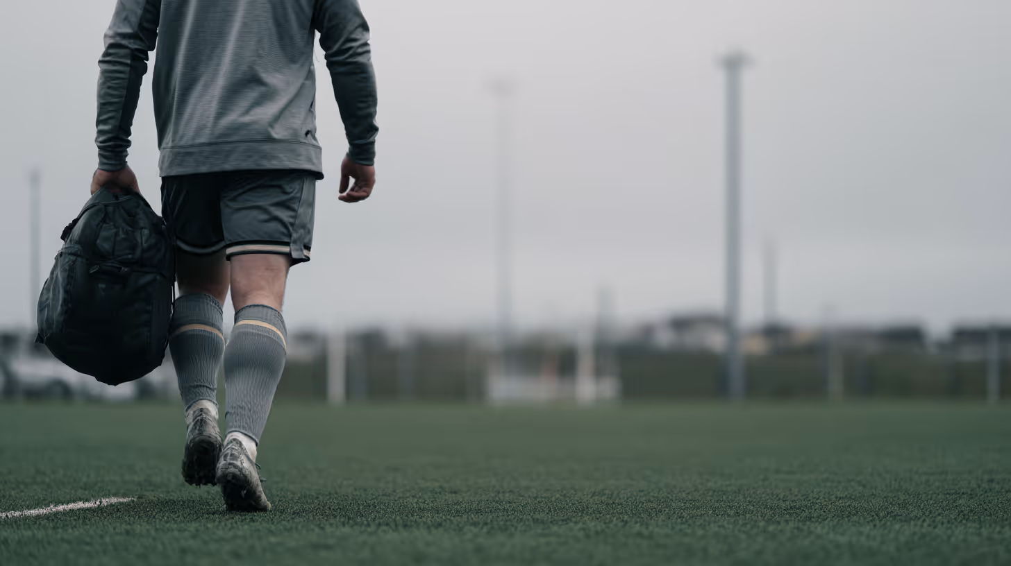Athlete in grey sportswear walking on a grassy field carrying a black duffel bag under overcast sky.