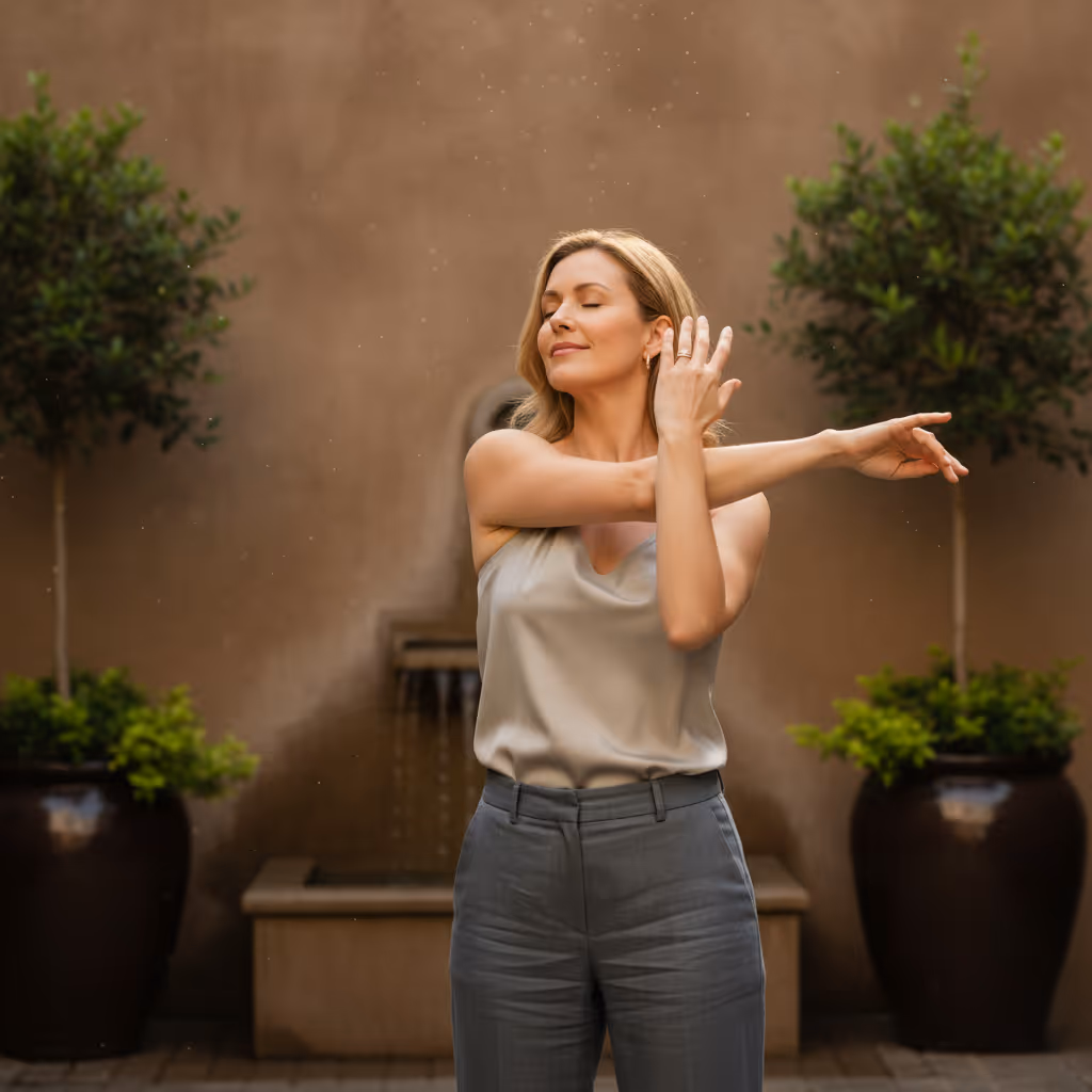Woman with closed eyes stretching her arms in a courtyard with potted plants and a fountain.