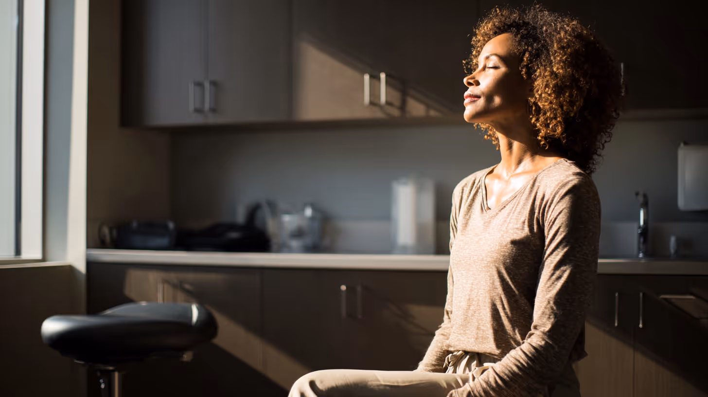 Woman with curly hair sitting peacefully with eyes closed in sunlit kitchen.