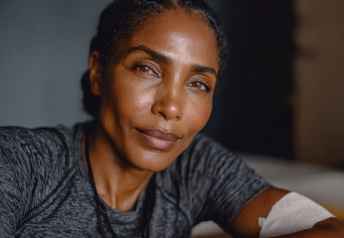 Close-up of a woman with brown eyes and braided hair wearing a gray shirt, resting with a bandage on her arm.