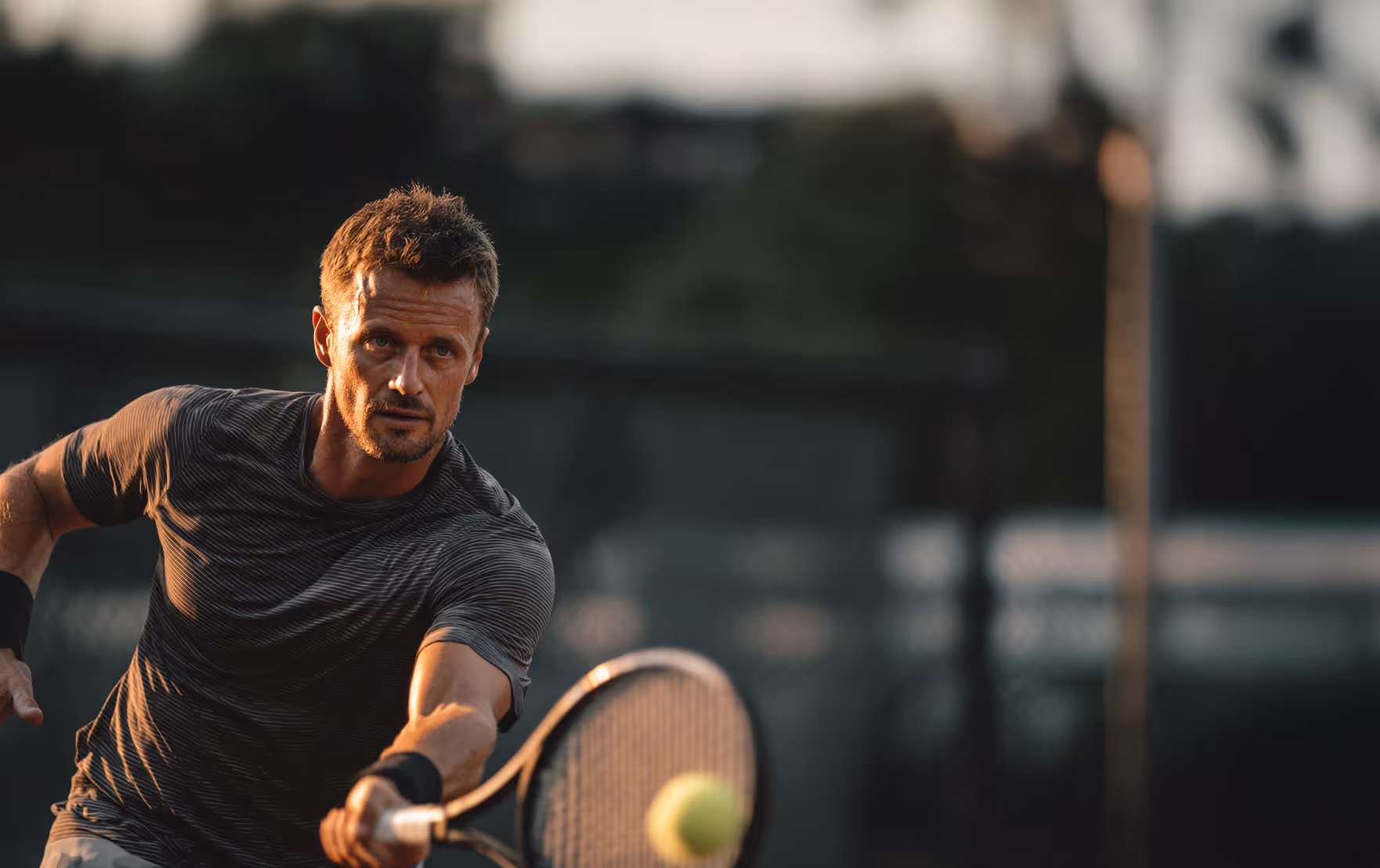 Focused male tennis player reaching to hit a yellow tennis ball with his racket on an outdoor court.