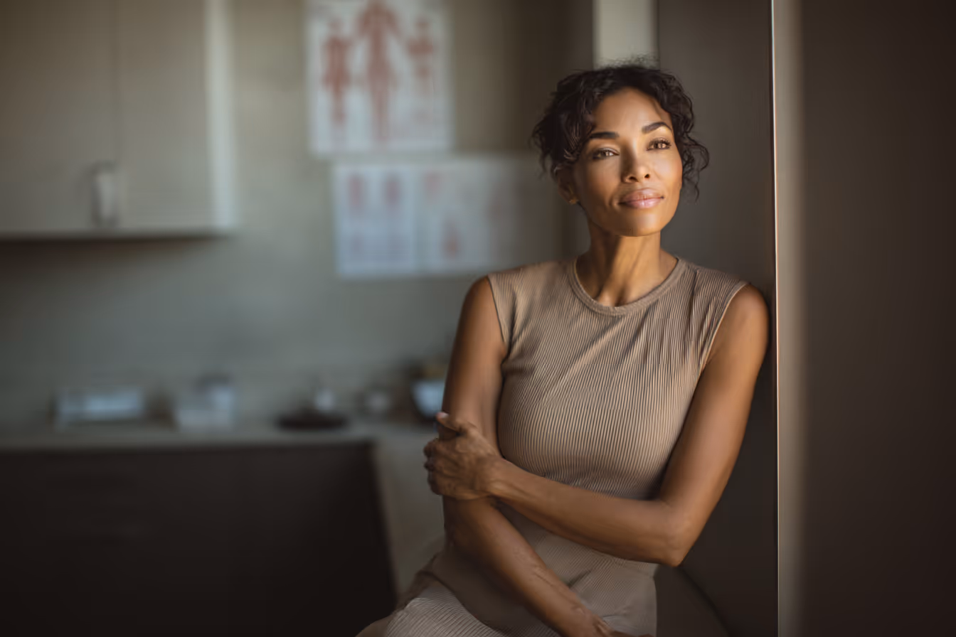 Thoughtful woman with curly hair in a sleeveless beige dress sitting by a window in a softly lit room with medical charts in the background.