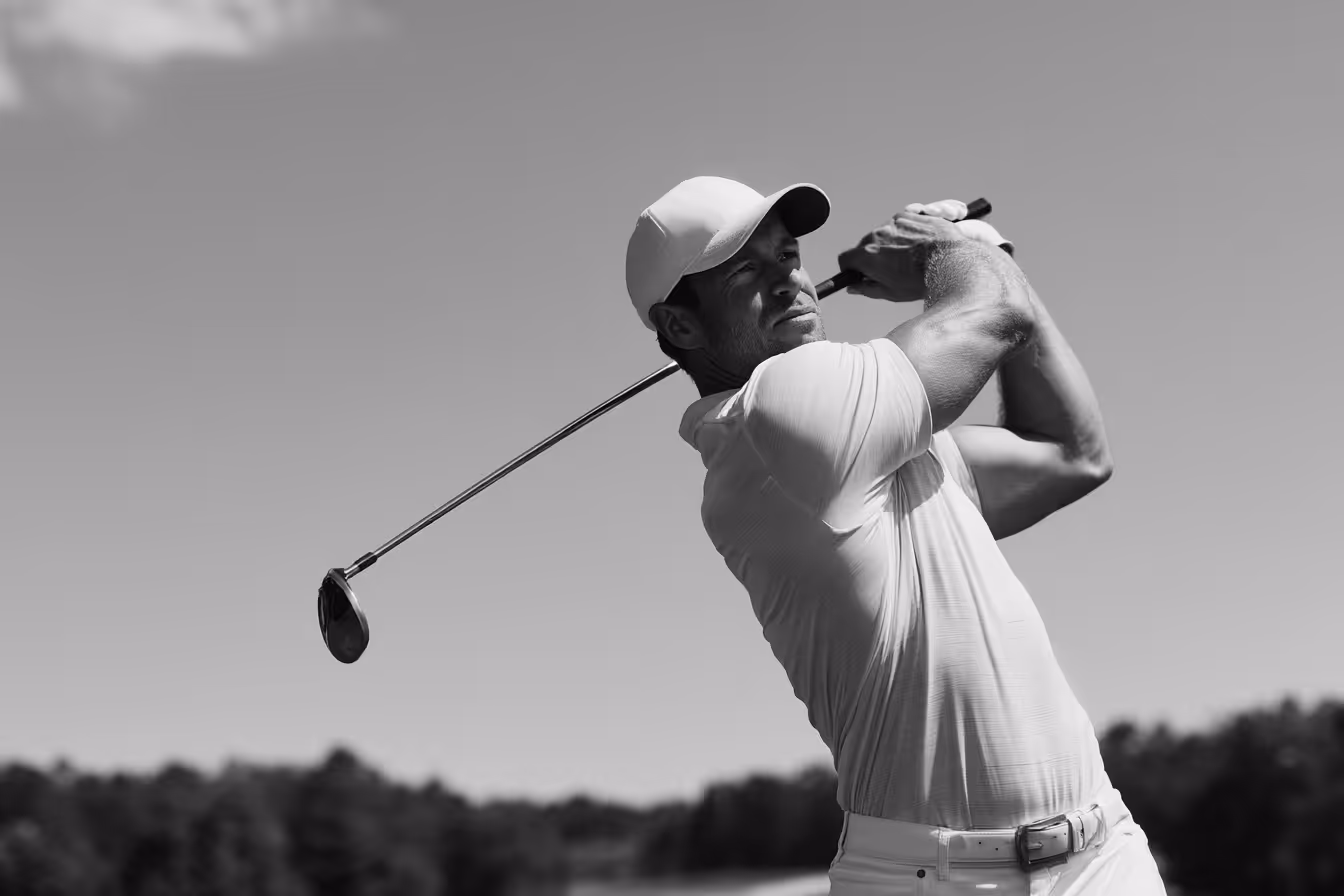 Man in a cap swinging a golf club on a sunny day with trees in the background.