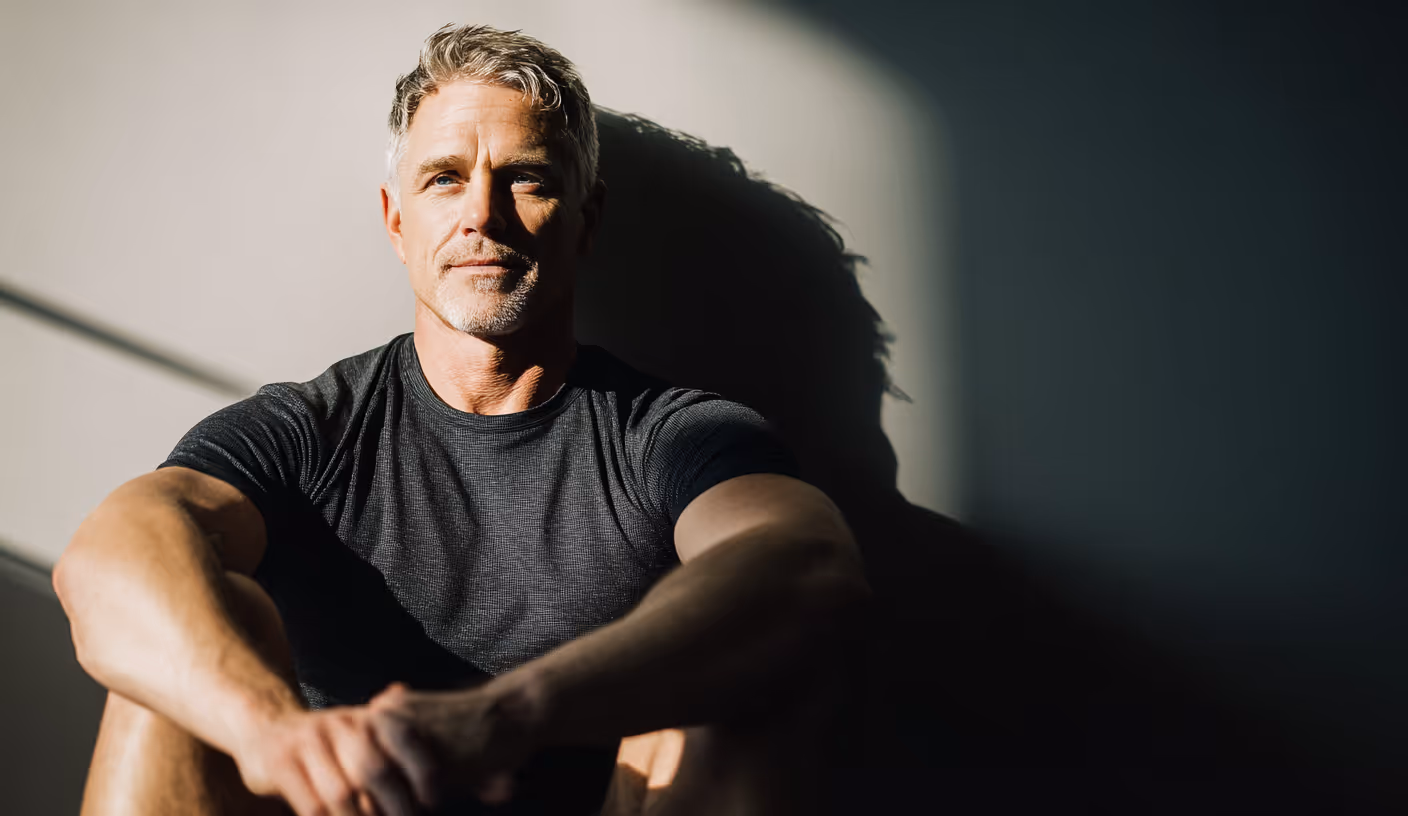 Middle-aged man with gray hair sitting against a wall with sunlight casting a shadow behind him.