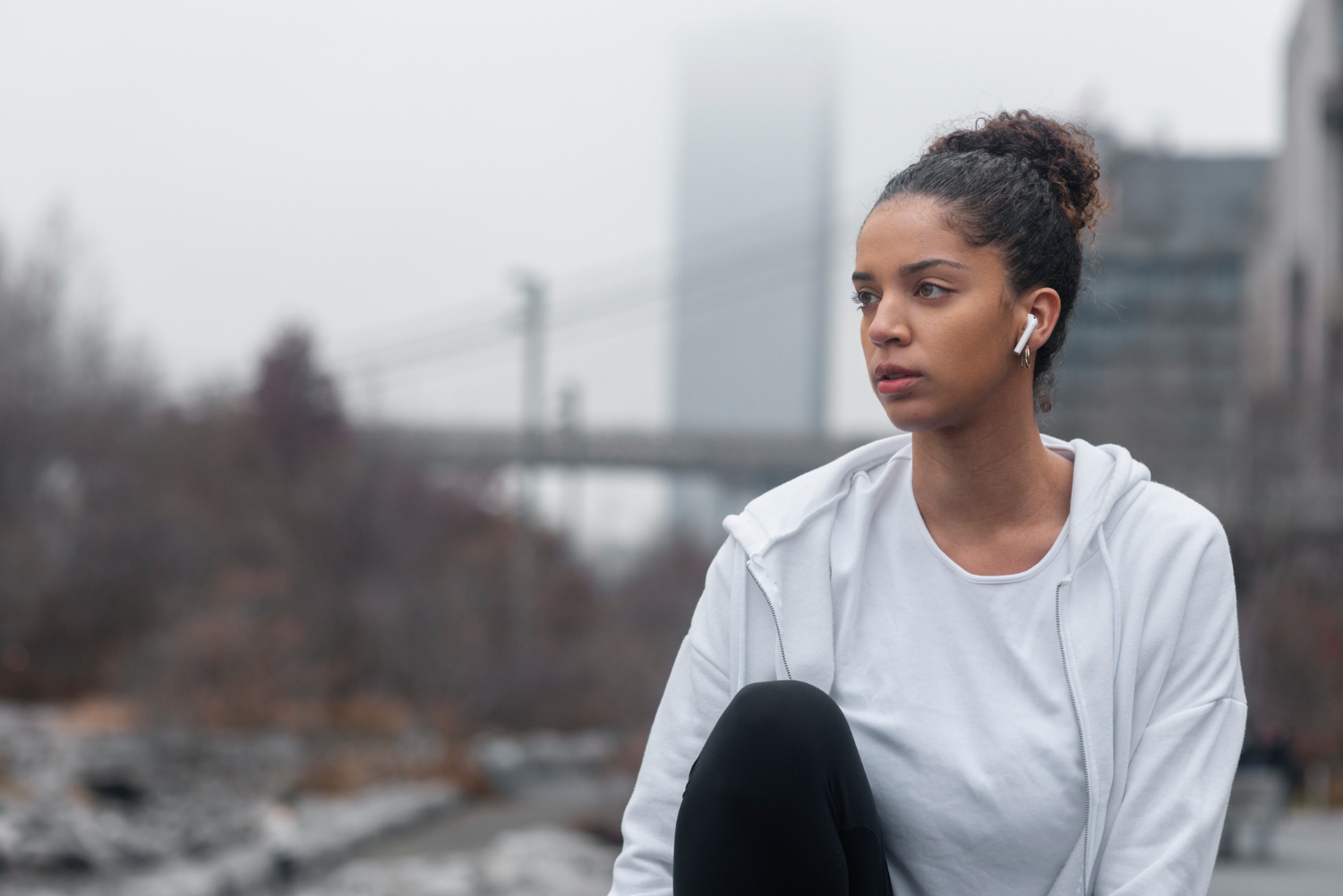 Young woman with curly hair in a bun wearing white earbuds and a white hoodie, sitting outdoors on a cloudy day.