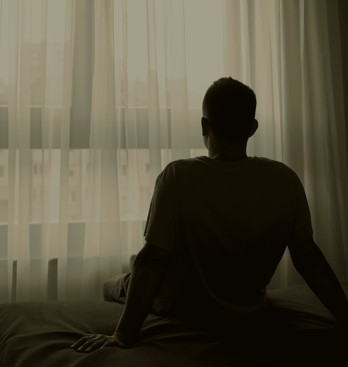 Silhouette of a person sitting on a bed looking out through sheer curtains at an urban landscape.