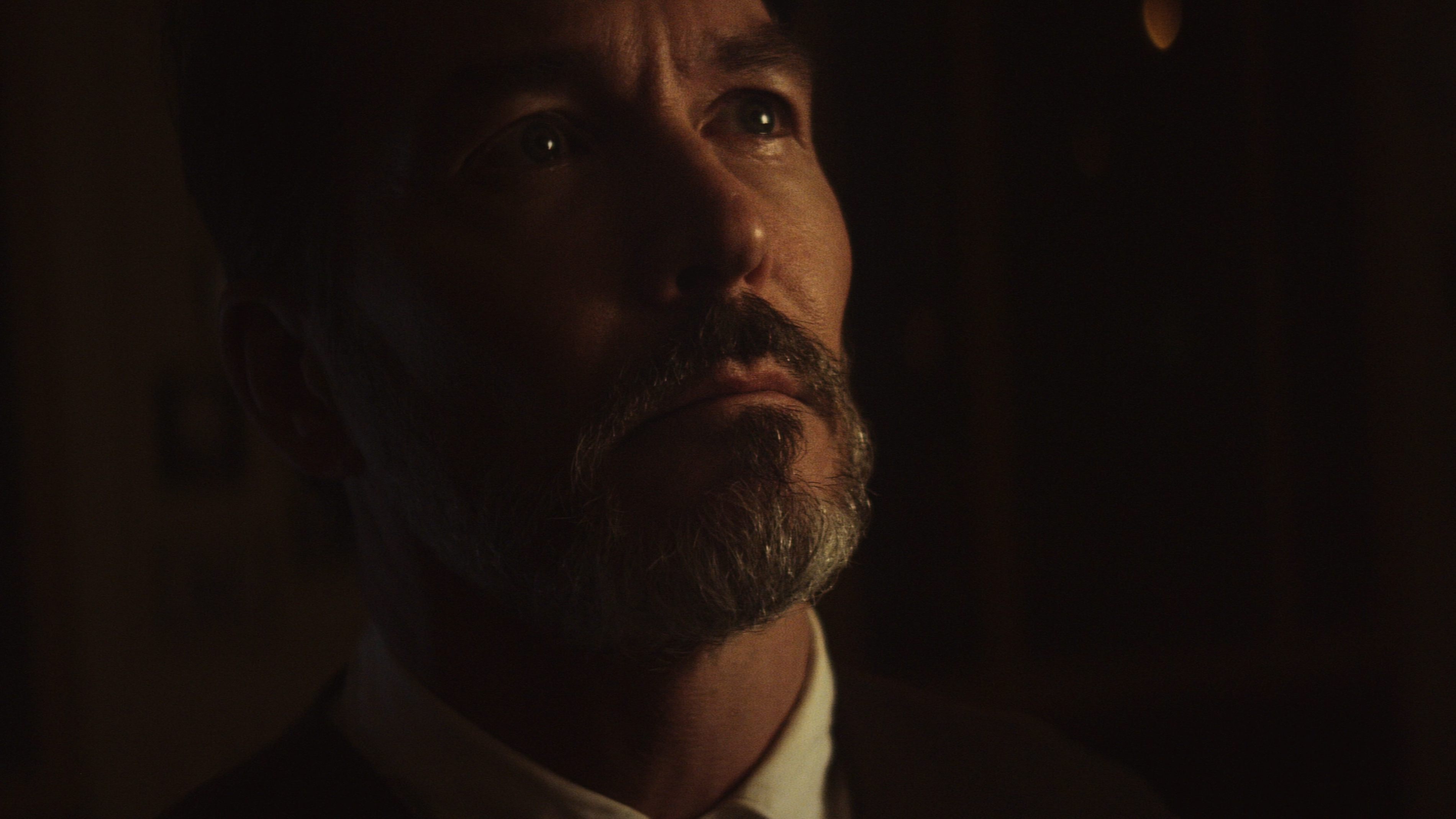 Close-up of a bearded man with a thoughtful expression, dimly lit against a dark background.