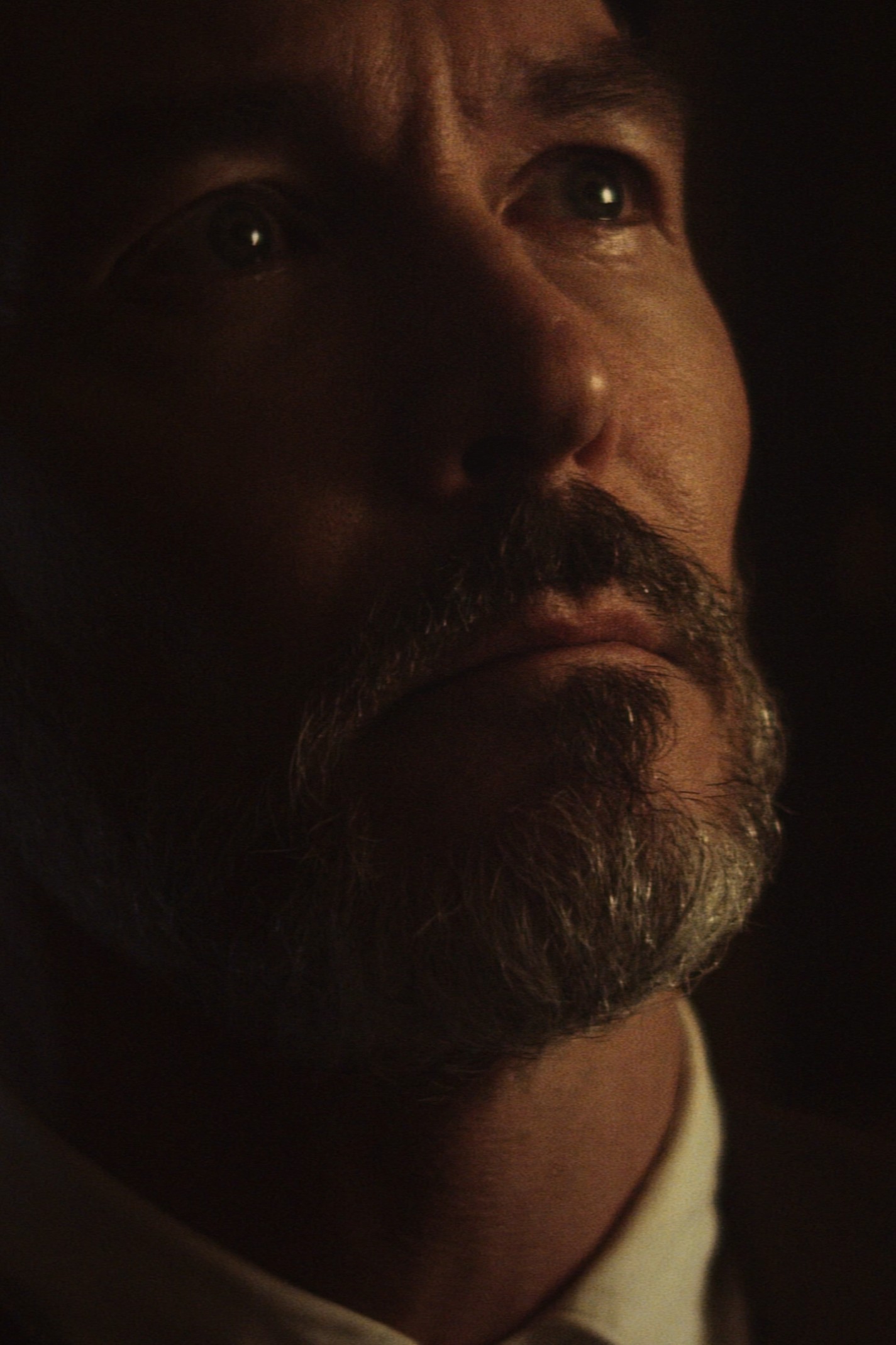 Close-up of a mature man with a mustache and beard looking upwards in low light.