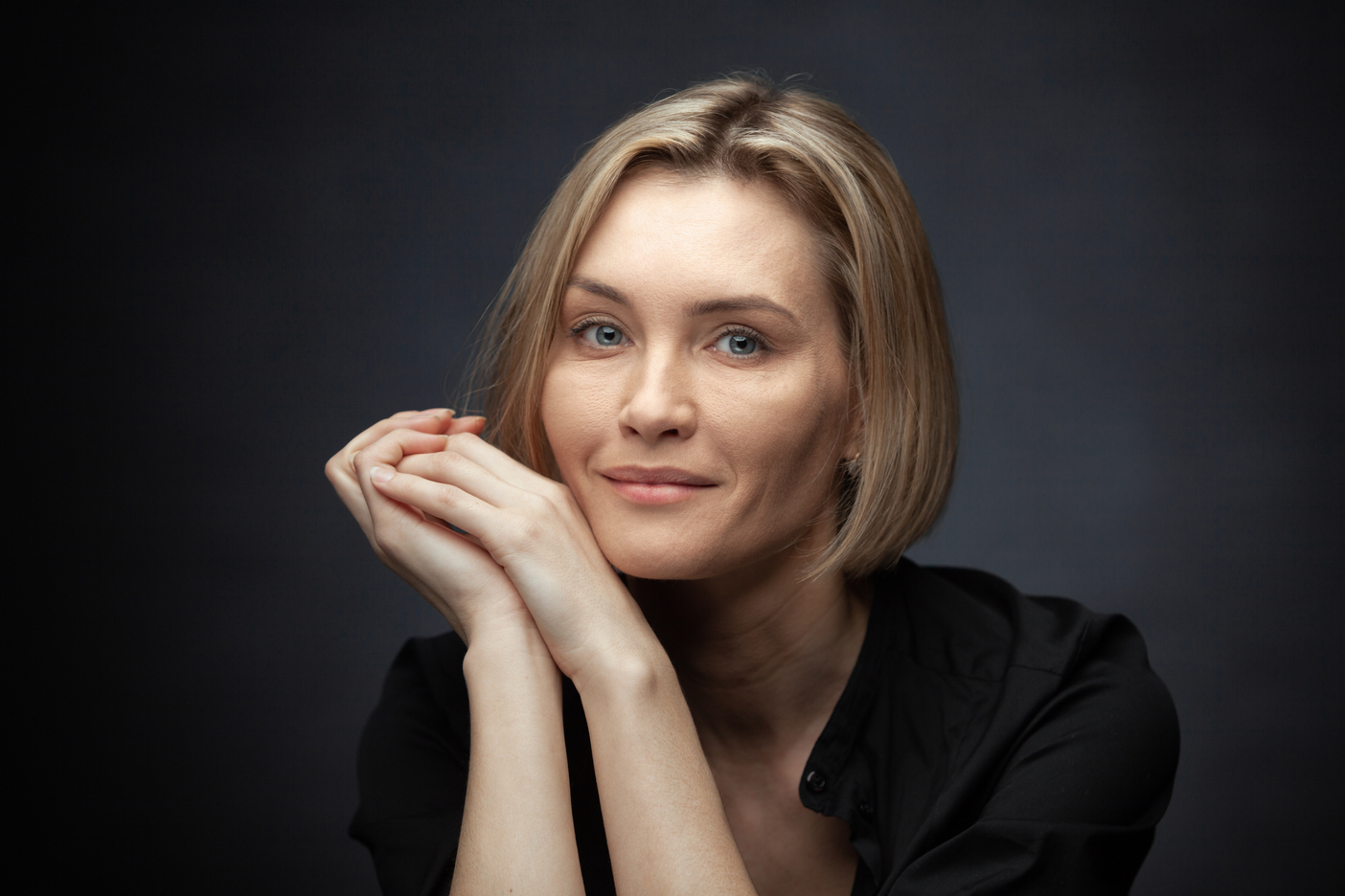Portrait of a blonde woman with blue eyes resting her chin on her folded hands against a dark background.