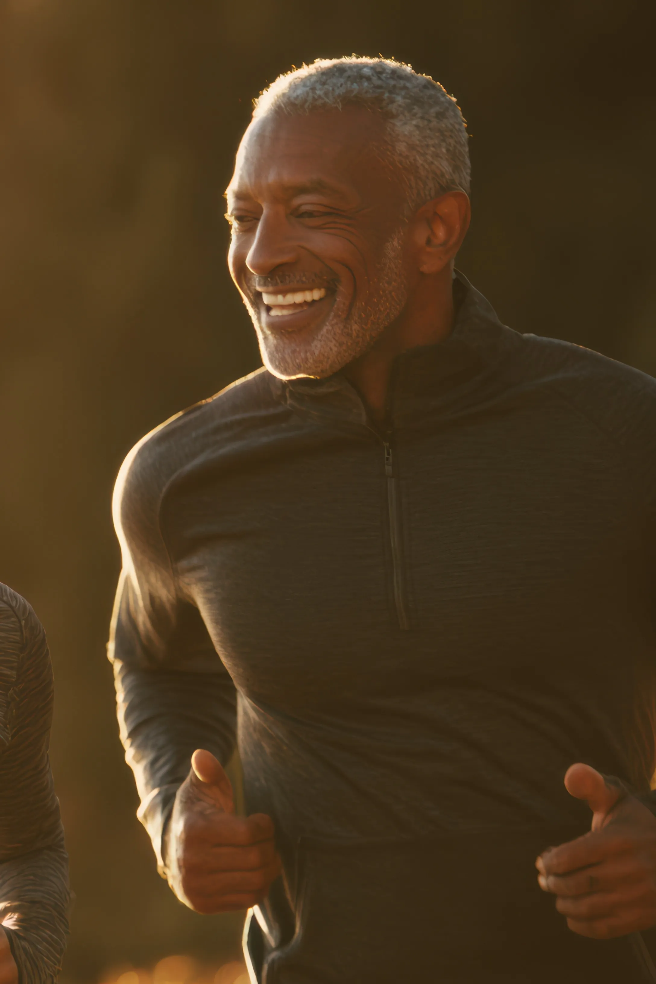 Smiling older man jogging outdoors in a dark long-sleeve zip-up shirt during golden hour.