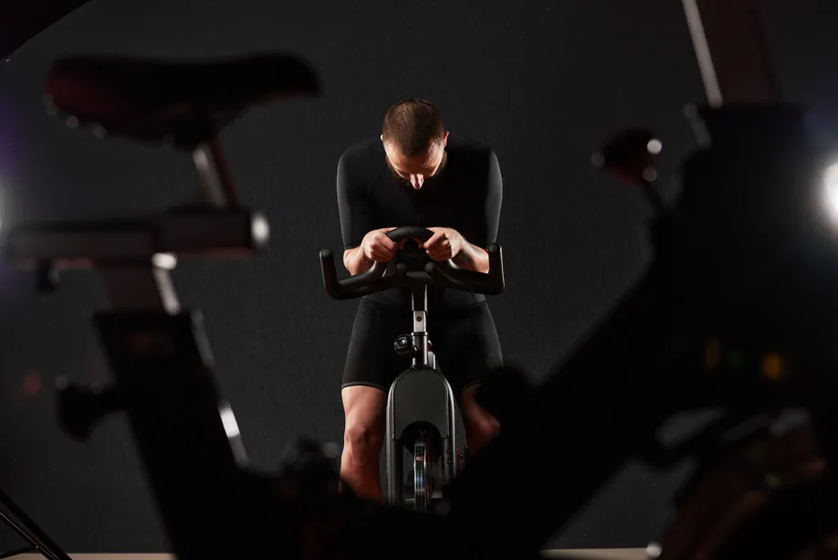 Man in black athletic wear leaning forward on an exercise bike against a dark background.