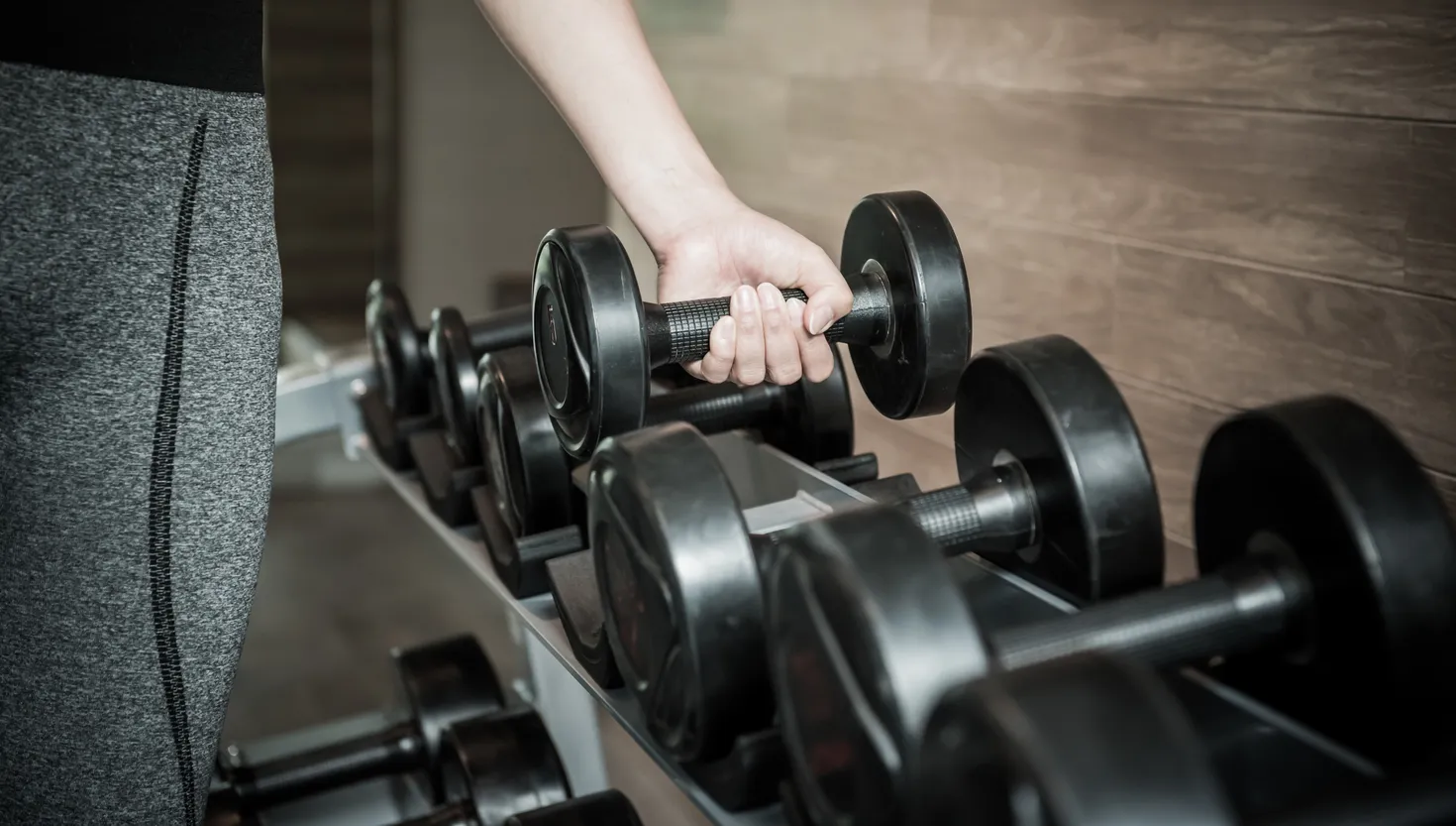 Person gripping a black dumbbell from a rack in a gym setting with wooden wall background.
