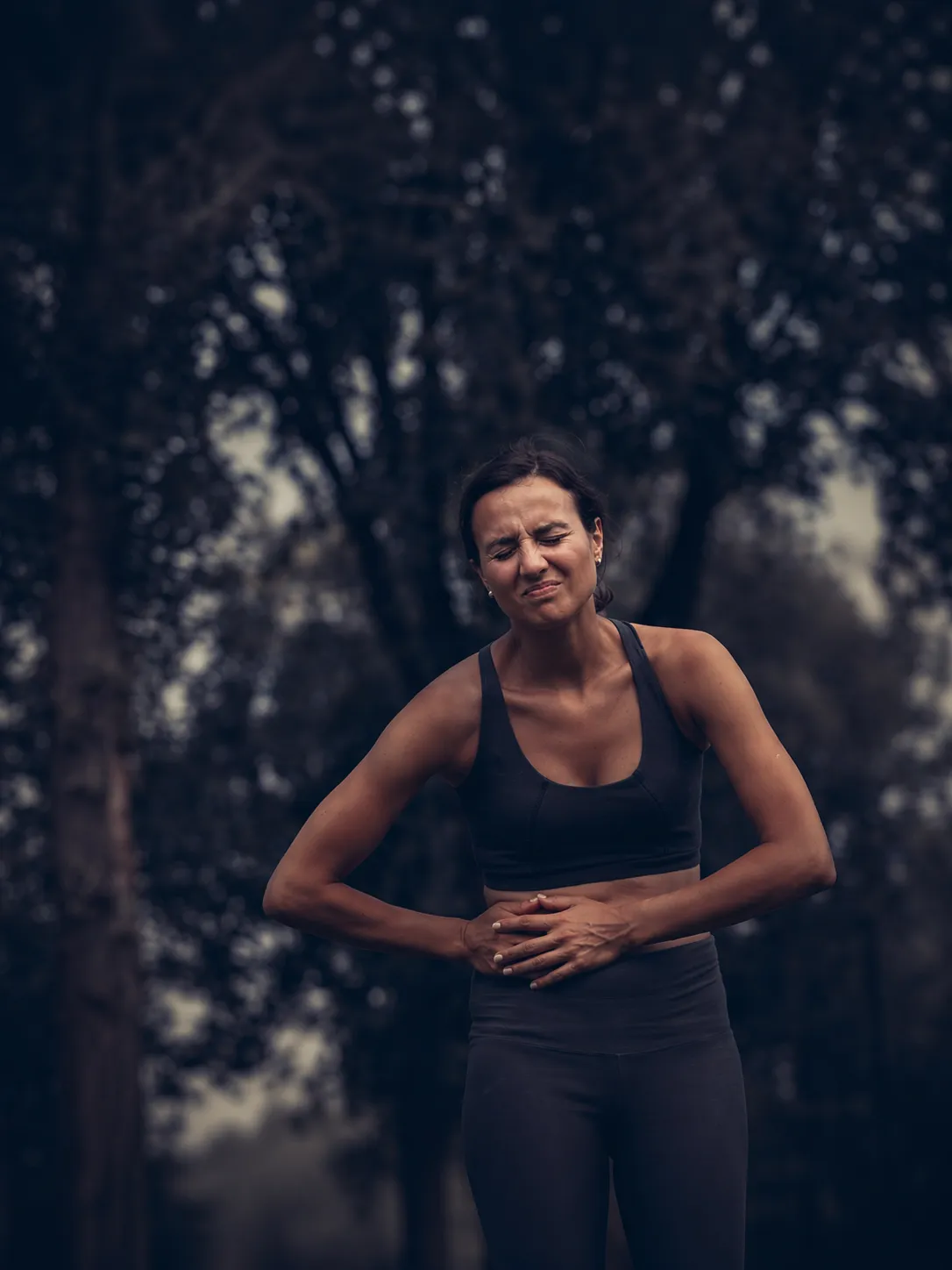 Woman in workout clothes outdoors clutching her abdomen in pain with eyes closed.