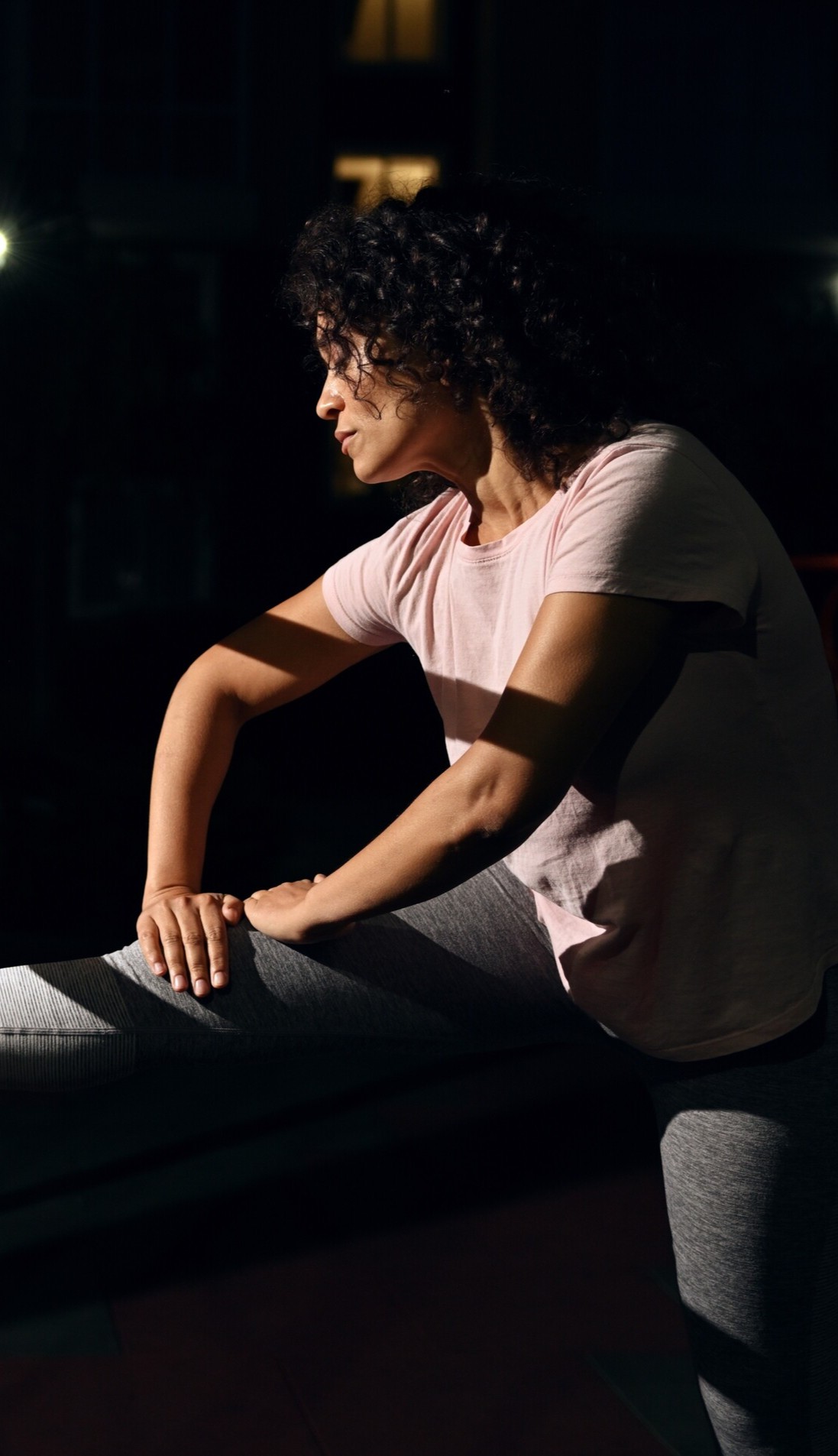 Woman with curly hair in a pink shirt and gray leggings stretching her leg indoors in low light.