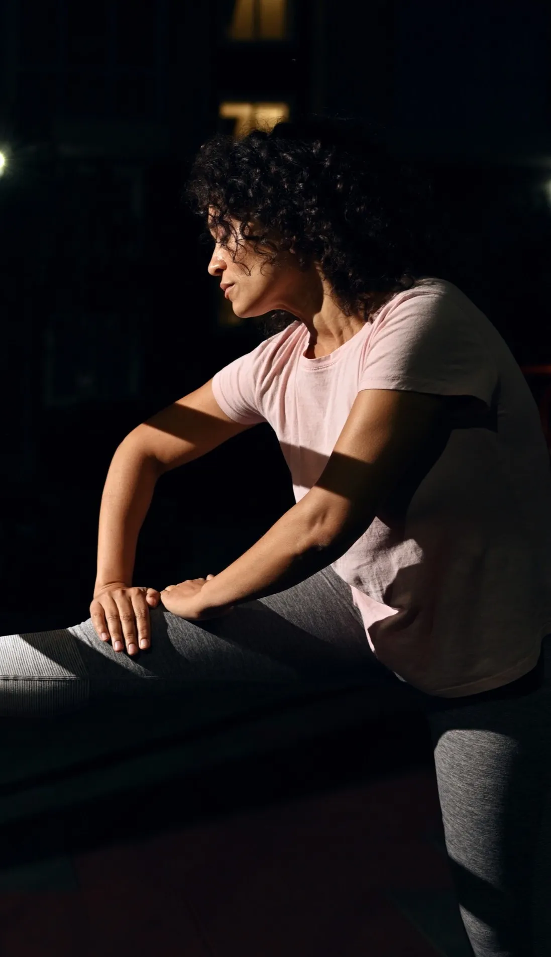 Woman with curly hair in a pink shirt and gray leggings stretching her leg indoors in low light.