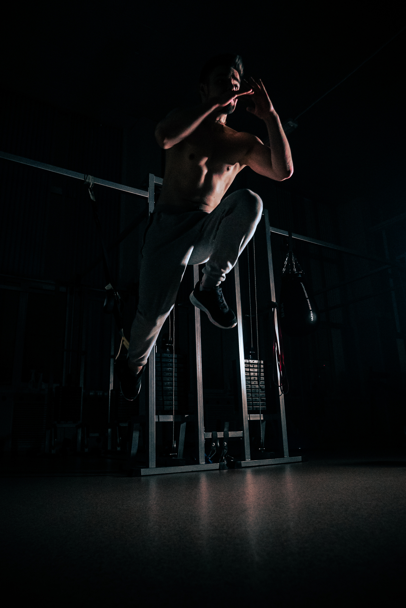 Shirtless man wearing gray sweatpants and black shoes jumping in a dimly lit gym.