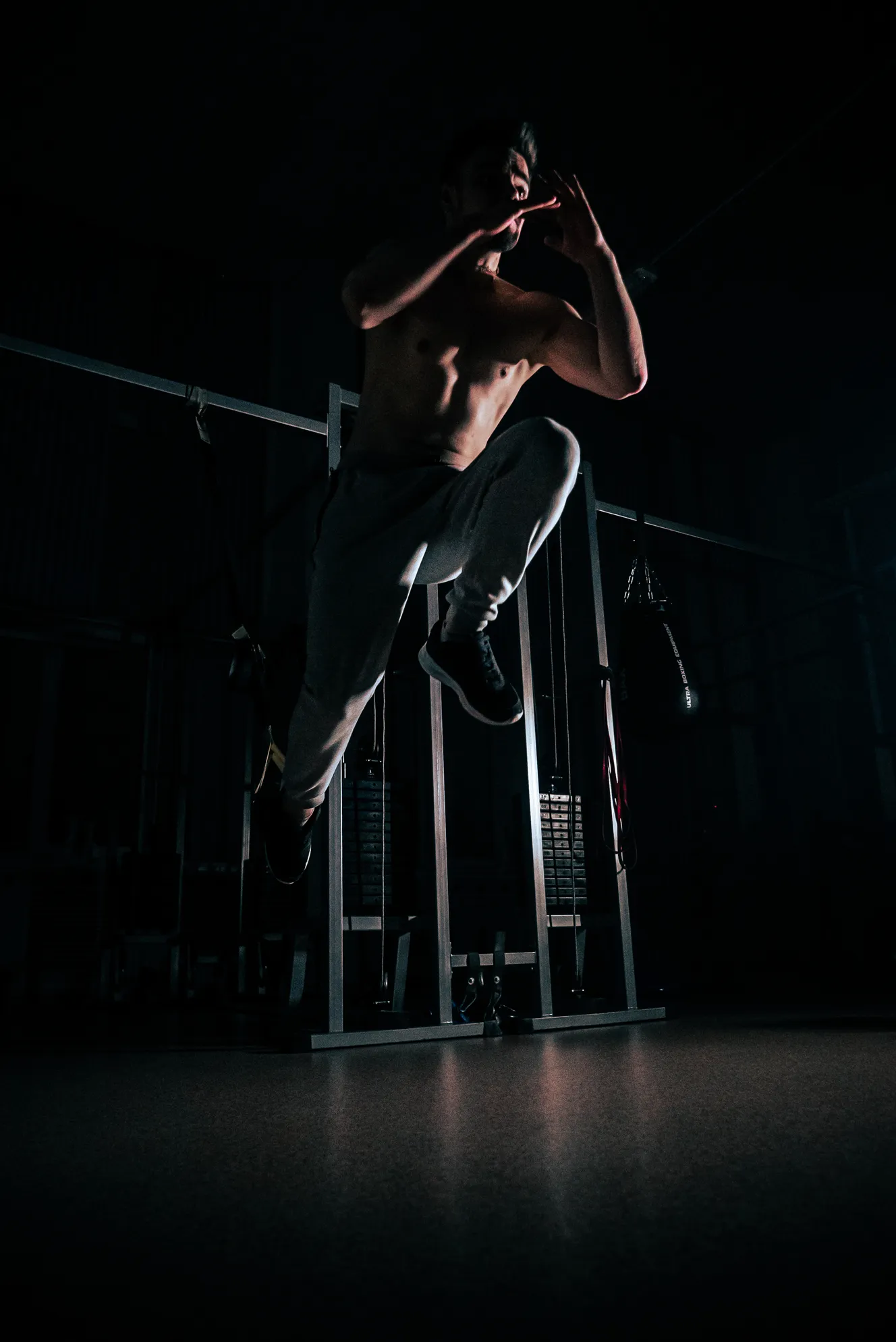 Shirtless man wearing gray sweatpants and black shoes jumping in a dimly lit gym.