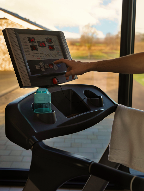Person's hand adjusting settings on a treadmill console next to a water bottle and towel.