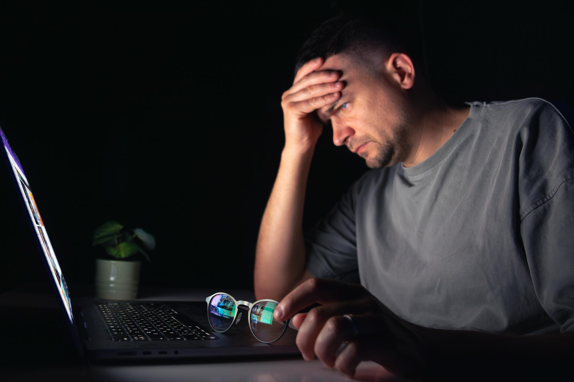 Person sitting at a laptop with a hand on their forehead, appearing fatigued from prolonged screen use, illustrating digital eye strain, stress, or screen-related fatigue.