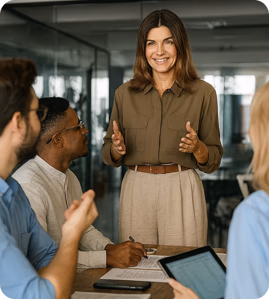 A smiling woman standing and speaking to three seated colleagues in a modern office meeting.