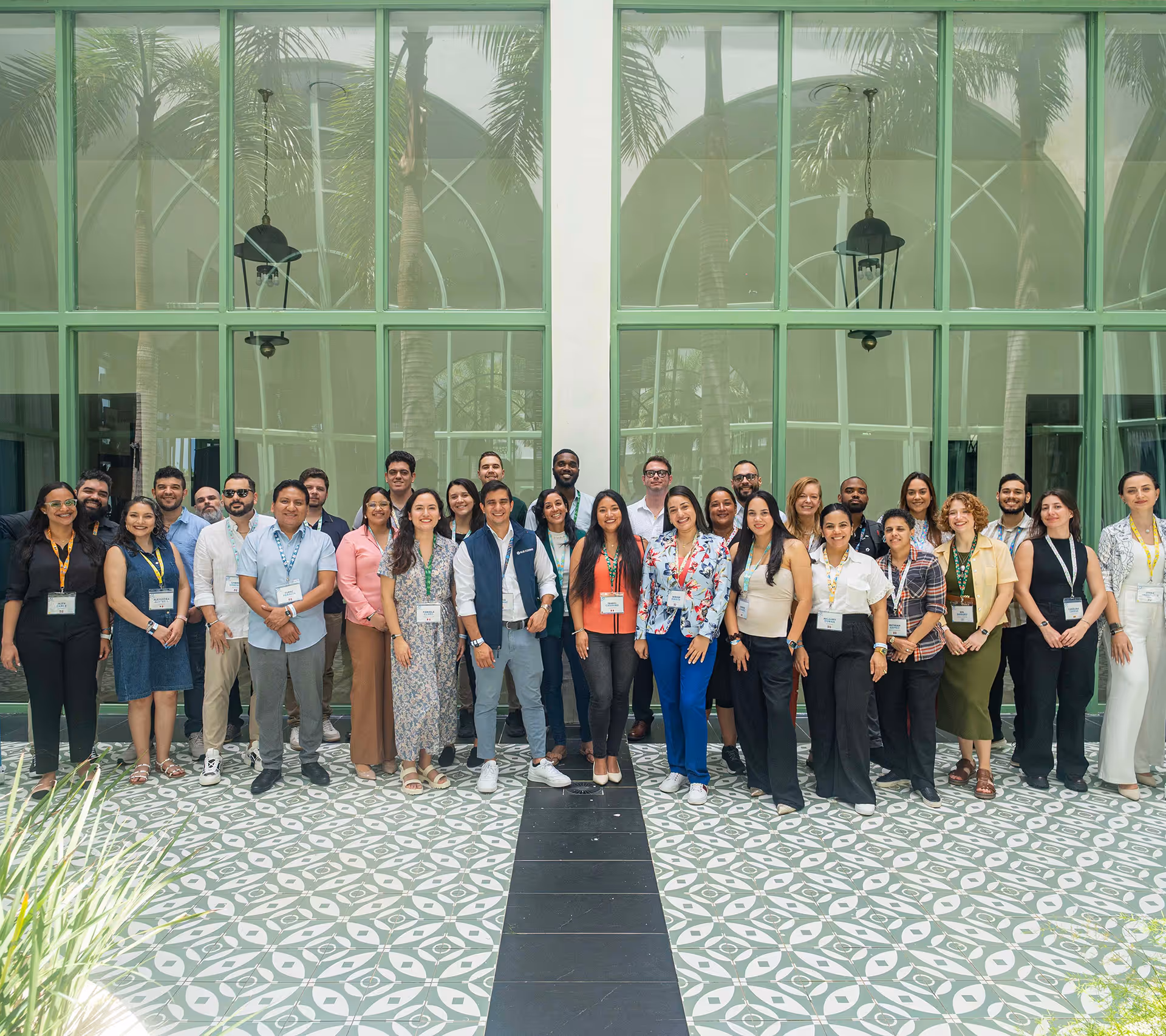 Group of diverse adults standing in a semi-formal setting, smiling in front of large green-framed windows and patterned floor tiles.
