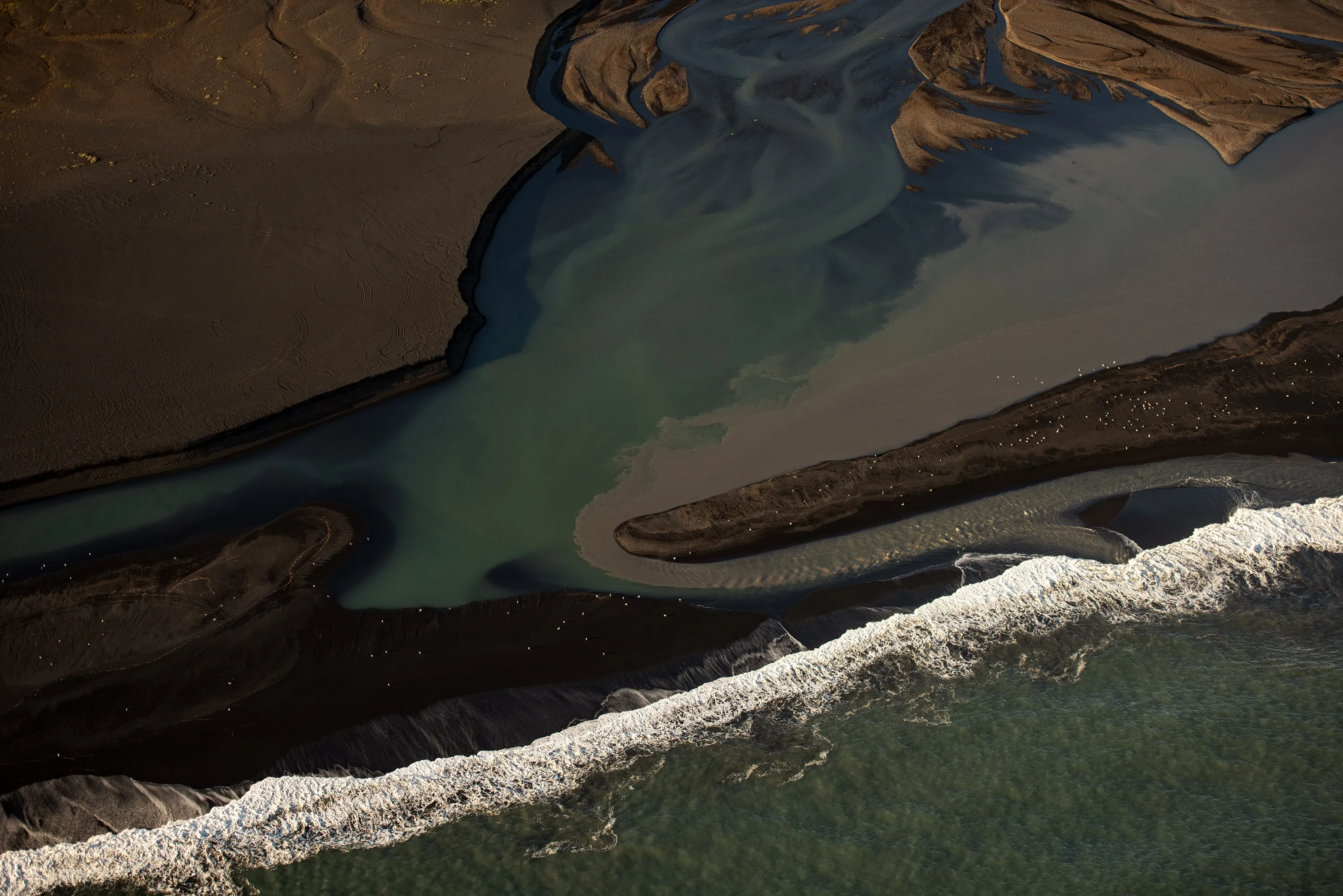 Aerial view of a river delta with dark sandbars, flowing into the ocean with white foamy waves.